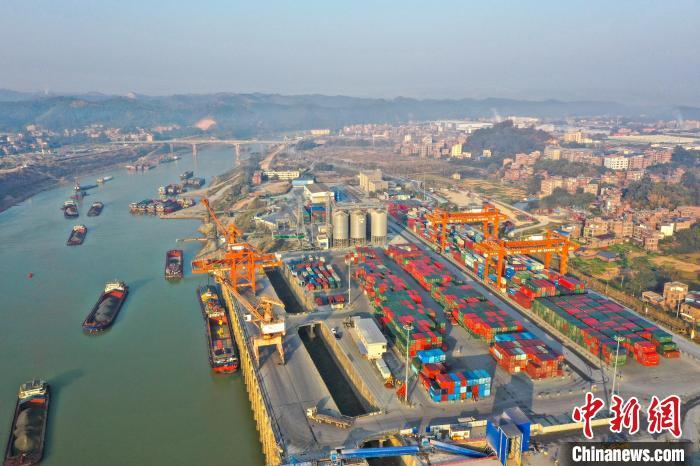 中国侨网1月4日,在广西梧州港大利口作业区,工作人员正在利用起吊设备吊装集装箱,货车在码头上来往穿梭,江面上货船有序航行,一派繁忙的景象。